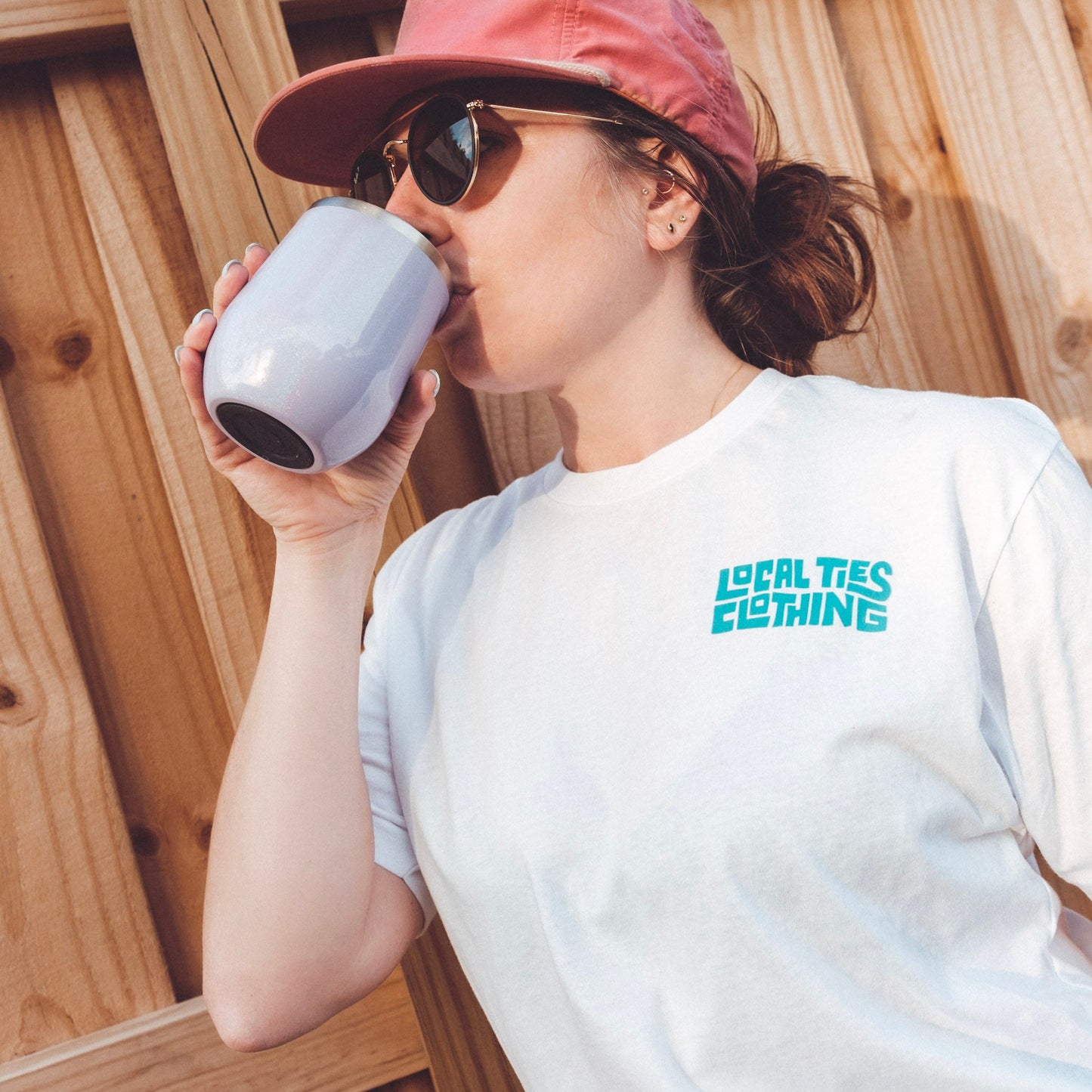 A woman wearing sunglasses and a Jubilee Tee from Local Ties Clothing, drinking from a mug.